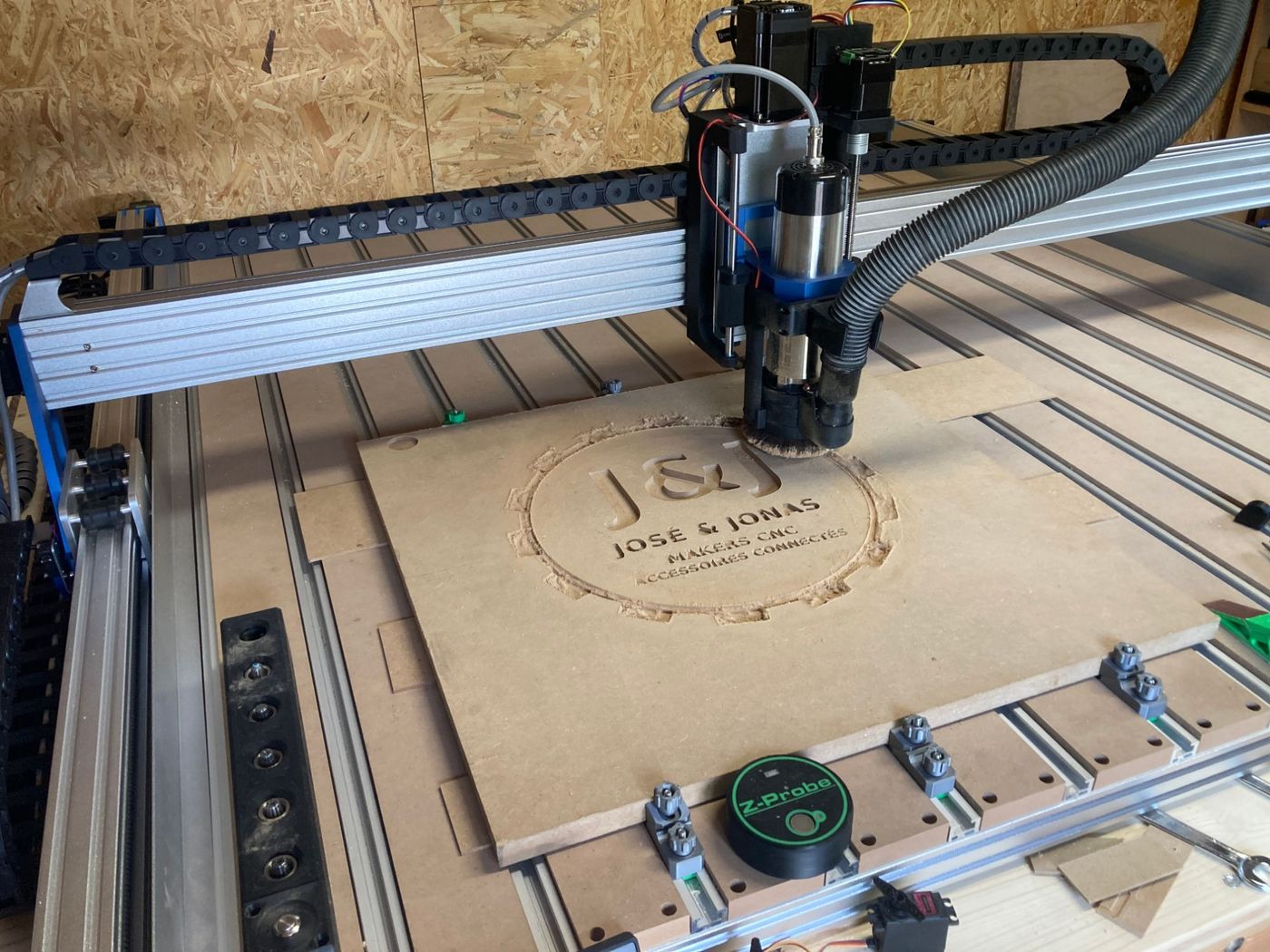 Fraiseuse CNC en train de graver le logo J&J dans une planche de MDF, avec la dust-shoe autour de la fraise et la sonde Z posée au bord du plateau